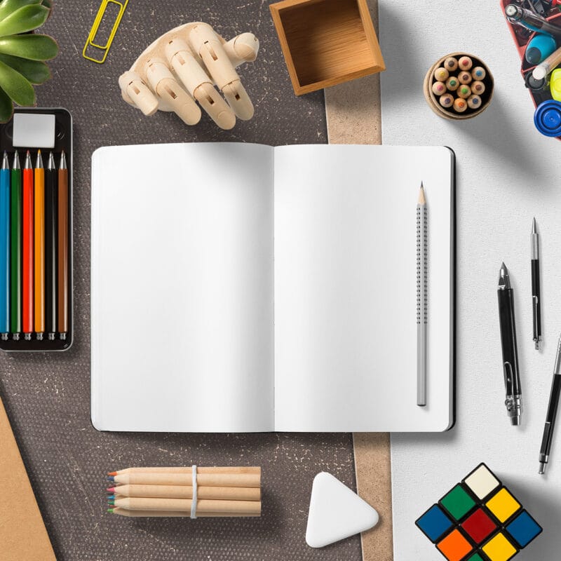 Overhead view of an open blank notebook on a desk surrounded by art and design tools, including colored pencils, pens, markers, a Rubik’s Cube, paper clips, and a small wooden mannequin hand.