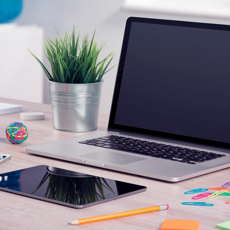 Laptop with a blank screen on a desk next to a potted plant, with a tablet, pencil, paper clips, sticky notes, and a small rubber band ball nearby.