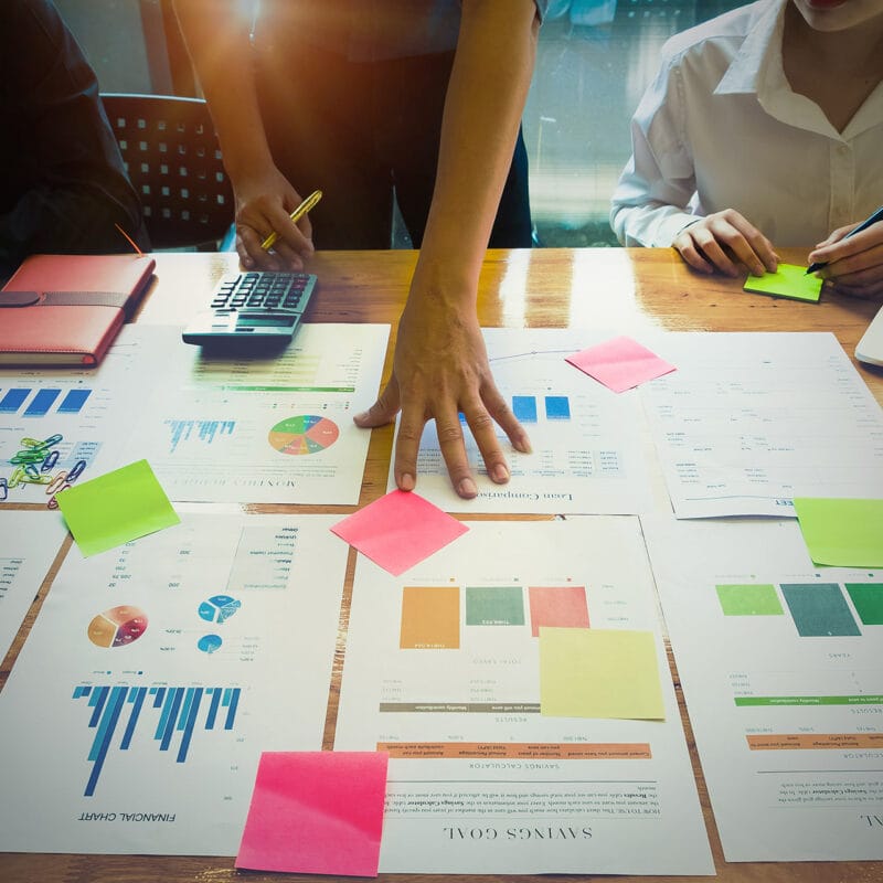 Three people at a meeting table review printed charts and reports, with colorful sticky notes on the pages, a calculator and notebooks nearby, and one person pointing at the documents.