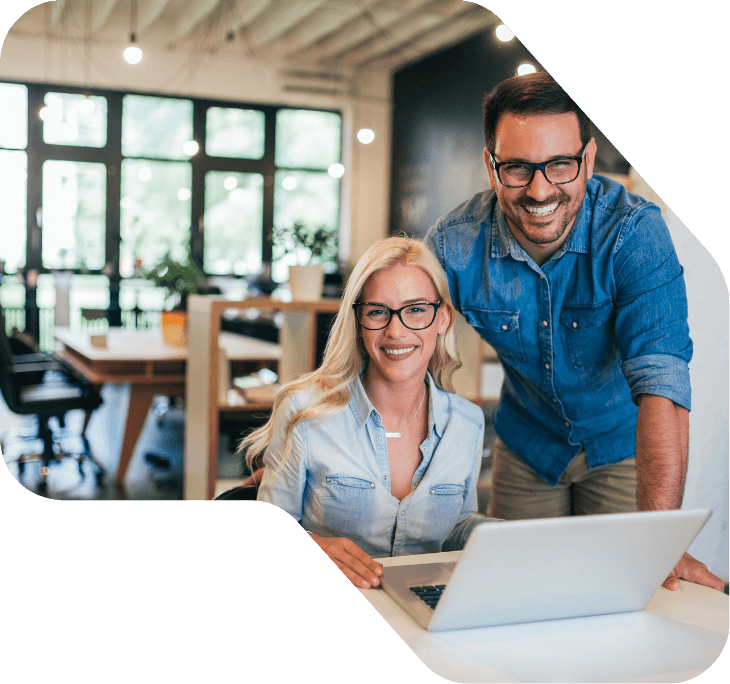 Smiling woman seated at a laptop while a smiling man leans in beside her in a bright, modern office with large windows.