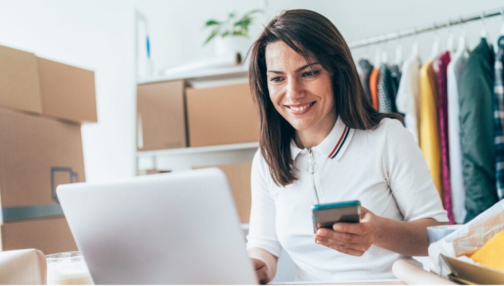 A woman smiles while using a laptop and holding a smartphone in a small shop workspace, with packing boxes and a rack of clothes in the background.