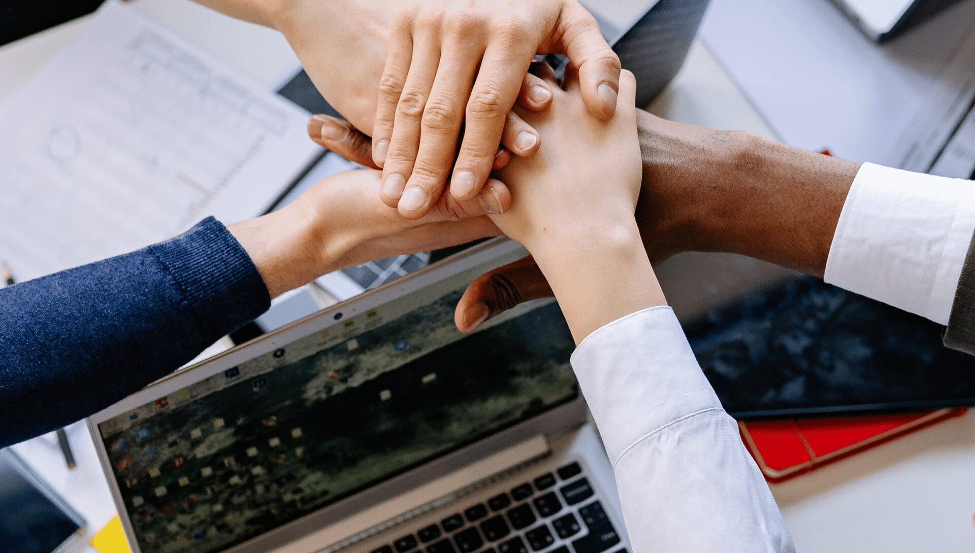 Close-up of five people stacking their hands together over a laptop on a desk, showing teamwork and collaboration.