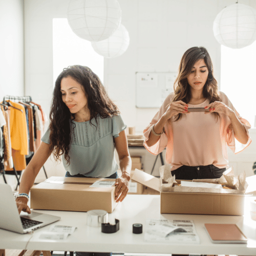 Two small business owners packing orders at a bright worktable, with shipping boxes, labels, and a laptop in a studio workspace.