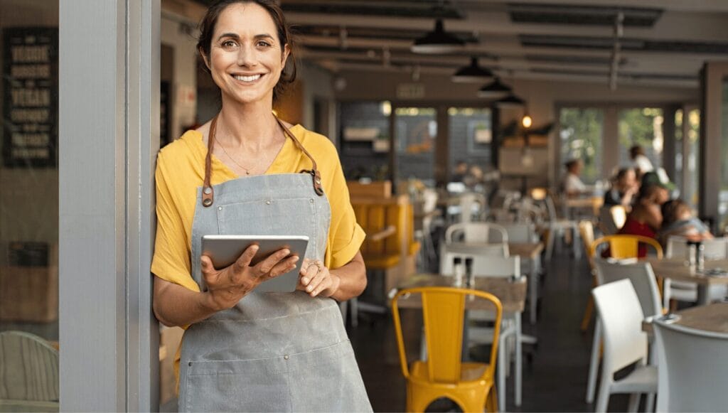 Smiling café owner in an apron stands at the entrance holding a tablet, with customers seated at tables in the background.