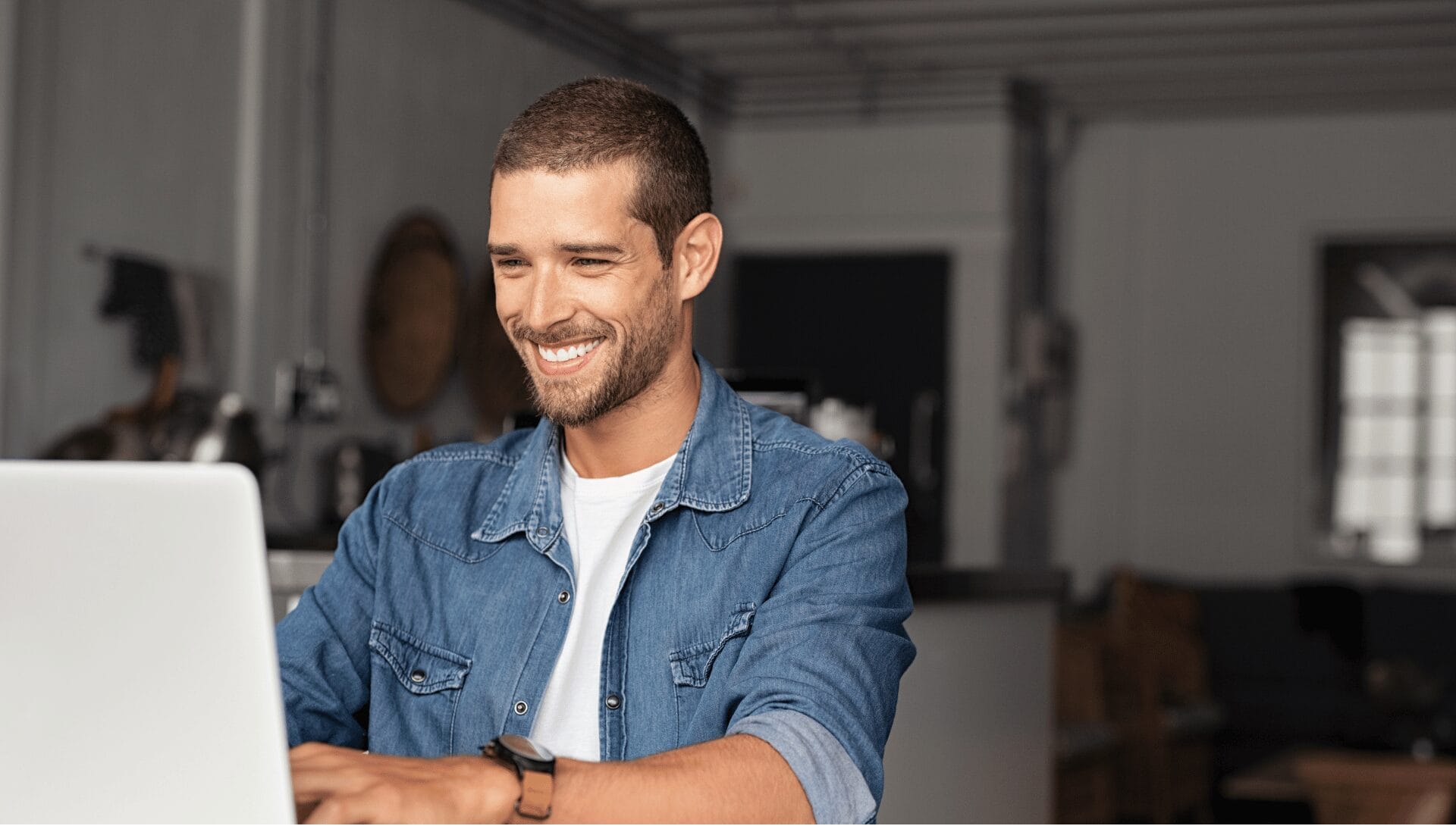 Smiling man works on a laptop at home, wearing a denim shirt, with a blurred kitchen in the background.