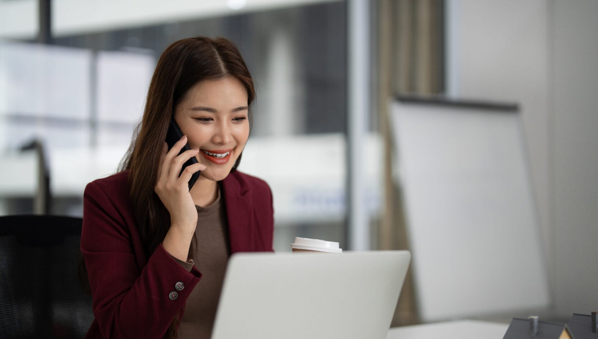 Smiling woman in an office talks on a phone while looking at her laptop.