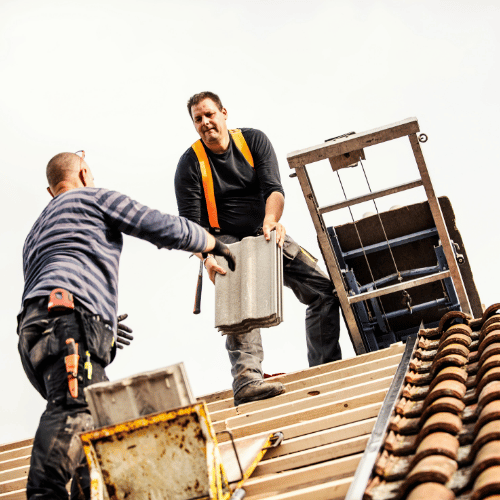 Two roofers working on a roof, with one handing roofing materials to the other beside a ladder and stacked tiles.
